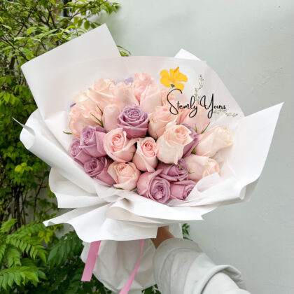 The bouquet features soft pastel-colored roses in shades of pale pink and lavender. The background shows green foliage and a plain light-colored wall