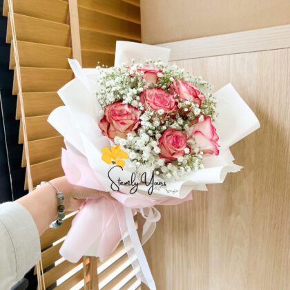Hand holding a bouquet of pink roses and white baby’s breath wrapped in pastel paper