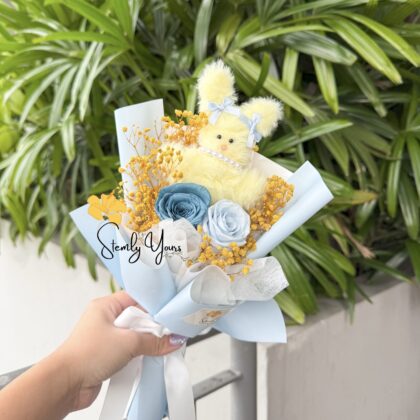 Mini bouquet with pastel blue roses, yellow baby’s breath, and a fluffy yellow bunny plush, wrapped in sky-blue paper with white ribbon