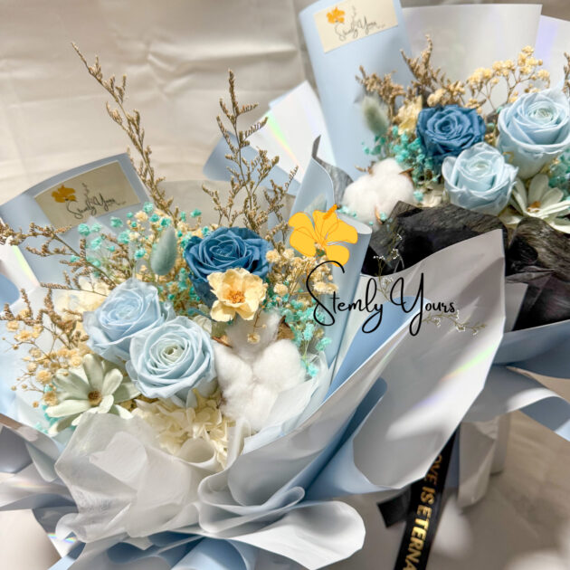 A dreamy bouquet featuring preserved roses in shades of sky blue and deep blue, accented with white cotton, baby's breath, dried foliage, and delicate flowers. The arrangement is wrapped in soft blue and white paper with a flowing, layered design, creating a serene and elegant presentation