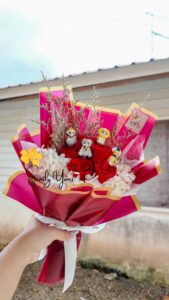 Hand holding a vibrant bouquet of red roses and white hydrangeas, wrapped in pink and maroon paper, topped with cute cartoon animal figurines
