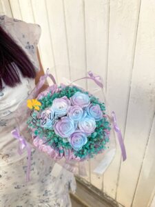 Person holding a pastel bouquet of lavender and baby blue roses with turquoise baby's breath, tied with lilac ribbons