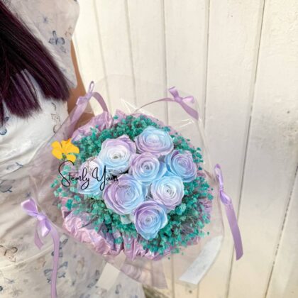 Person holding a pastel bouquet of lavender and baby blue roses with turquoise baby's breath, tied with lilac ribbons