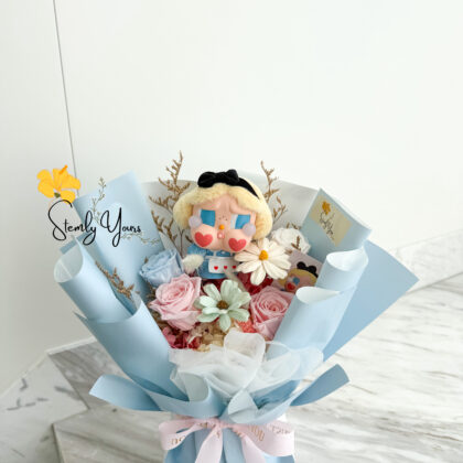 A pastel-themed bouquet featuring preserved roses in soft pink and baby blue, accent flowers, and a central doll-like figure with blonde hair, blue eyeshadow, heart-shaped cheeks, and a black bow headband. The bouquet is wrapped in light blue paper and white tulle, tied with a pink ribbon labeled “JUST FOR YOU.” The arrangement is displayed on a marble surface with a clean white background. The brand "Stemly Yours" is subtly watermarked on the left.