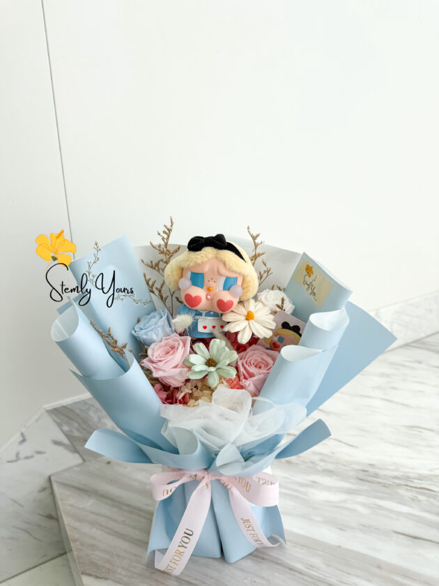 A pastel-themed bouquet featuring preserved roses in soft pink and baby blue, accent flowers, and a central doll-like figure with blonde hair, blue eyeshadow, heart-shaped cheeks, and a black bow headband. The bouquet is wrapped in light blue paper and white tulle, tied with a pink ribbon labeled “JUST FOR YOU.” The arrangement is displayed on a marble surface with a clean white background. The brand "Stemly Yours" is subtly watermarked on the left.