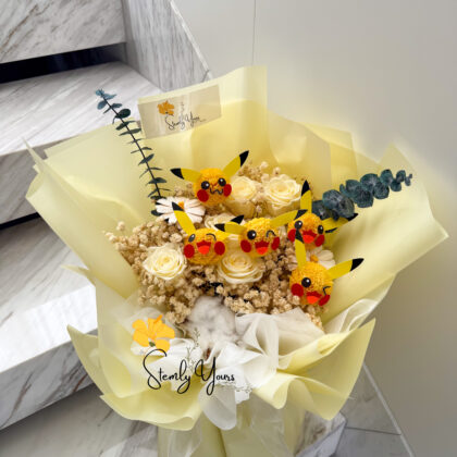 A cheerful bouquet wrapped in soft yellow paper, featuring preserved white roses, dried florals, cotton, eucalyptus, and multiple handcrafted Pikachu faces made from yellow pom-poms with red cheeks and black-tipped ears. The arrangement is topped with a “Stemly Yours” tag and sits on a marble staircase.