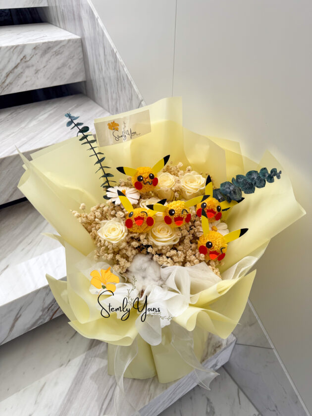 A cheerful bouquet wrapped in soft yellow paper, featuring preserved white roses, dried florals, cotton, eucalyptus, and multiple handcrafted Pikachu faces made from yellow pom-poms with red cheeks and black-tipped ears. The arrangement is topped with a “Stemly Yours” tag and sits on a marble staircase.