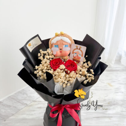 A hand-held bouquet wrapped in sleek black paper and tied with a red satin ribbon, featuring a teary-eyed doll with a fuzzy brown hat and white earmuffs at the top. The arrangement includes two preserved red roses surrounded by fluffy white baby’s breath, creating a bold and expressive floral presentation. A "Stemly Yours" tag is tucked into the bouquet.