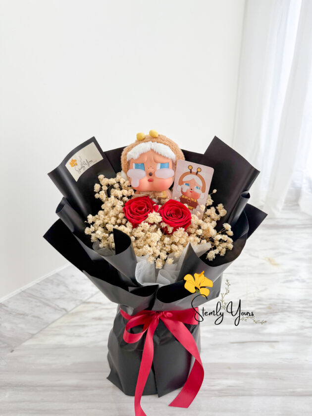 A hand-held bouquet wrapped in sleek black paper and tied with a red satin ribbon, featuring a teary-eyed doll with a fuzzy brown hat and white earmuffs at the top. The arrangement includes two preserved red roses surrounded by fluffy white baby’s breath, creating a bold and expressive floral presentation. A "Stemly Yours" tag is tucked into the bouquet.