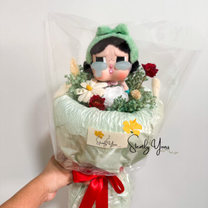 A hand-held bouquet wrapped in soft mint green paper and tied with a red satin ribbon, featuring a doll-like figurine with a green headband, pink cheeks, and large teardrops under its eyes. The bouquet includes small preserved flowers such as white daisies, red roses, and green foliage, all enclosed in a clear plastic wrapping. A "Stemly Yours" tag is attached to the bouquet
