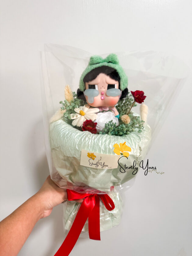 A hand-held bouquet wrapped in soft mint green paper and tied with a red satin ribbon, featuring a doll-like figurine with a green headband, pink cheeks, and large teardrops under its eyes. The bouquet includes small preserved flowers such as white daisies, red roses, and green foliage, all enclosed in a clear plastic wrapping. A "Stemly Yours" tag is attached to the bouquet